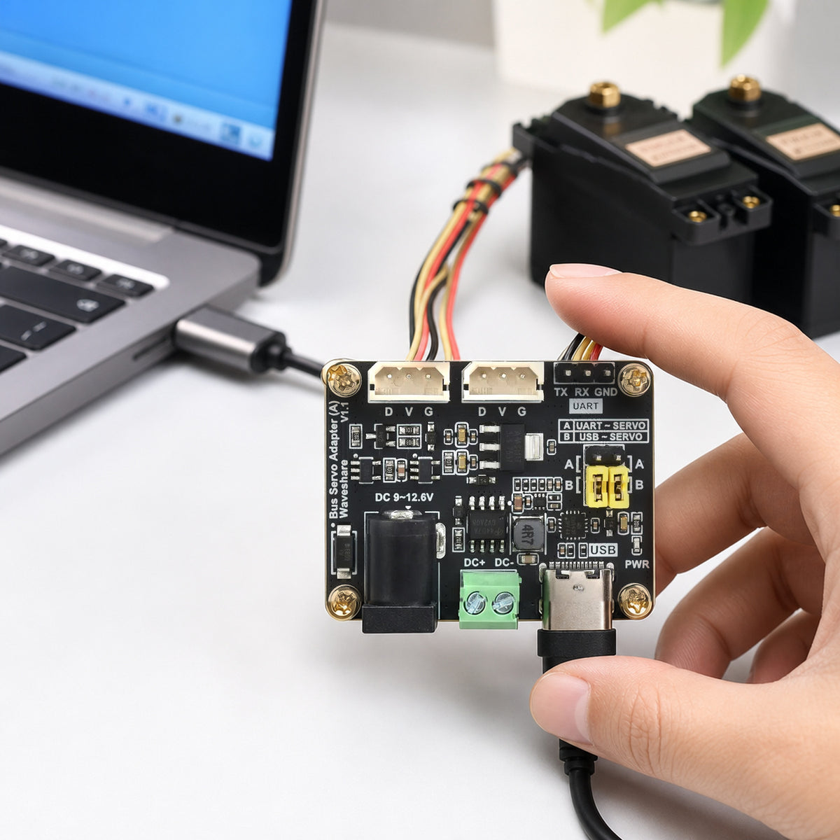Waveshare bus servo adapter board – the board on a desk wired by USB-C to a laptop and by a 3-pin cable to two black bus servos