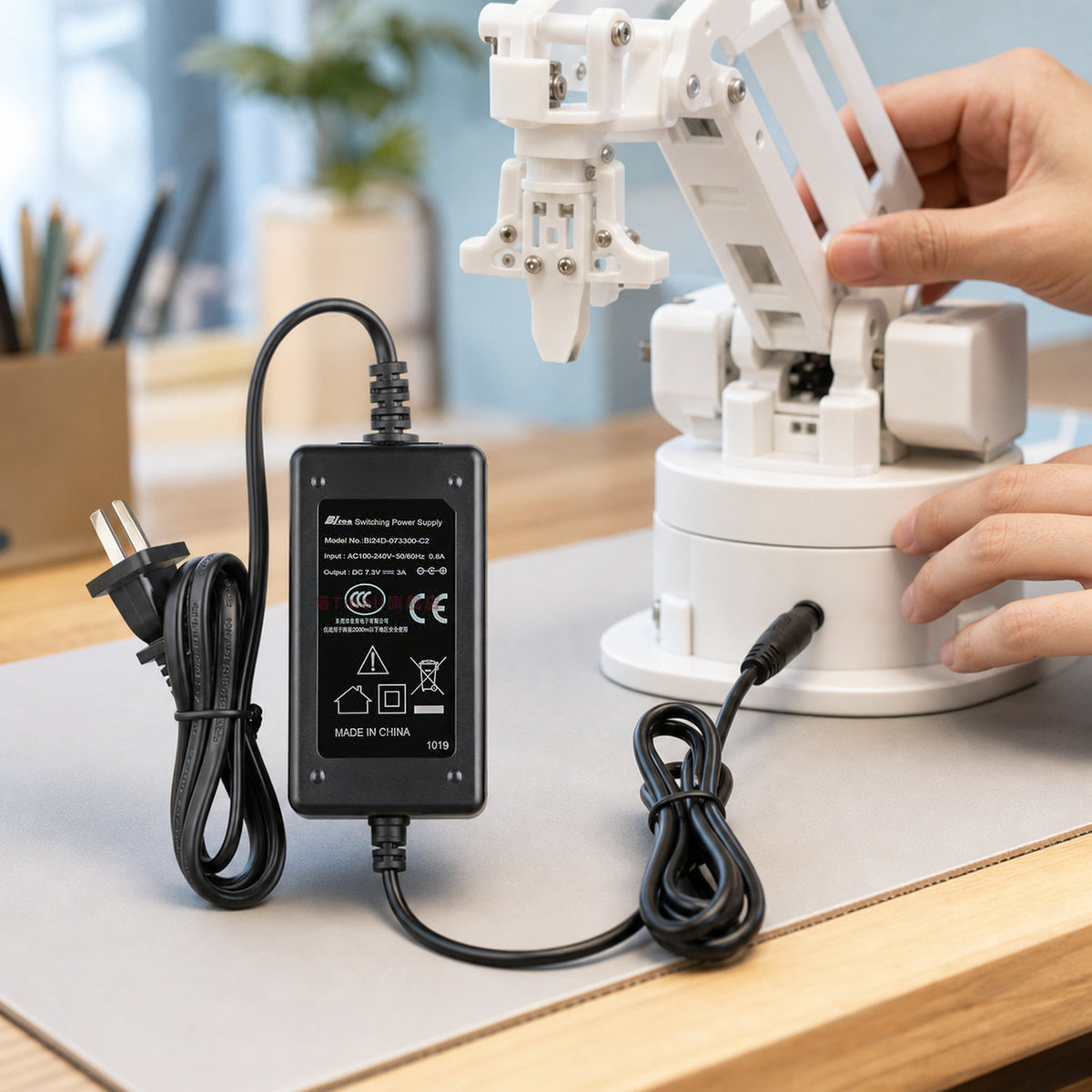 7.4 V servo power adapter – the adapter powering a small white 3D-printed desktop robot arm on a desk